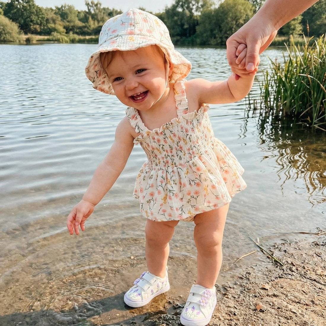 petite fille les pieds dans l'eau avec sa barboteuse et son chapeau qui tient papa et rit
